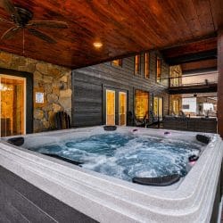 Hot tub on a covered patio beside a stone wall, with the rear of the cabin behind it