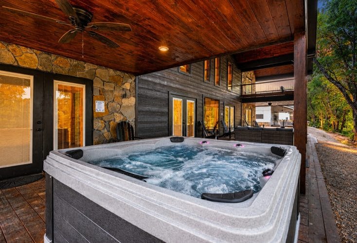 Hot tub on a covered patio beside a stone wall, with the rear of the cabin behind it
