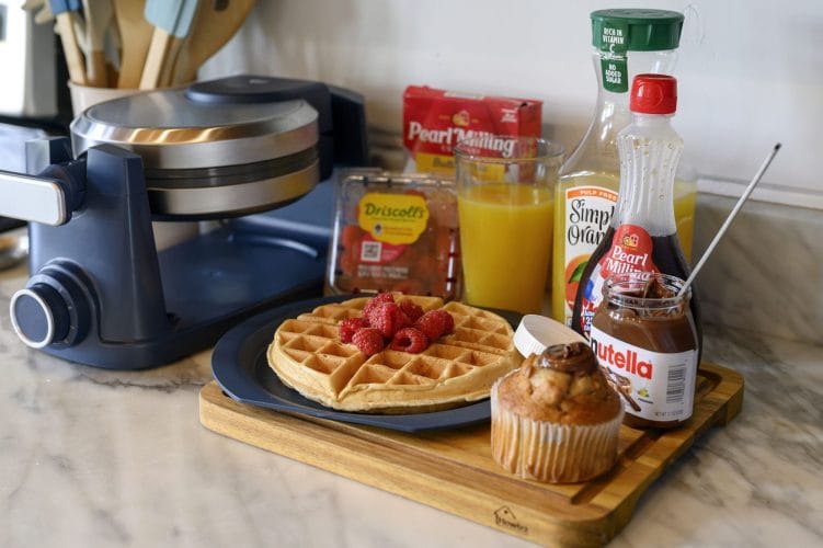 Waffle with berries, orange juice, syrup and a muffin on a tray beside a waffle maker
