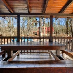Large wooden table with built-in benches on a covered deck, with woods beyond the railing