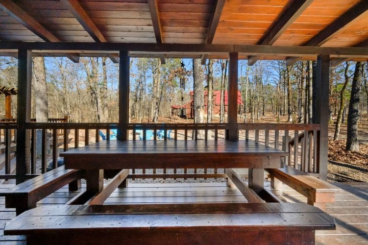 Large wooden table with built-in benches on a covered deck, with woods beyond the railing