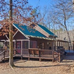 Front of the cabin with a green metal roof, porch with steps, and a deck, among bare trees