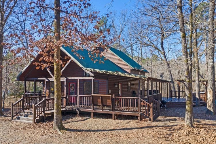 Front of the cabin with a green metal roof, porch with steps, and a deck, among bare trees
