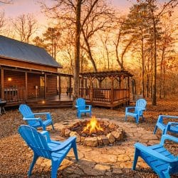 Round stone fire pit with a fire burning and blue Adirondack chairs around it, the cabin and a pergola behind, at sunset