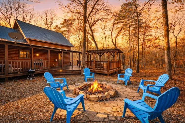Round stone fire pit with a fire burning and blue Adirondack chairs around it, the cabin and a pergola behind, at sunset