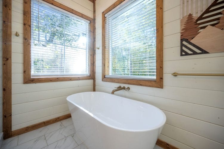 Freestanding tub under two windows beside mountain-pattern wall art