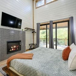 Same bedroom with a gas fireplace in a gray tile wall under the TV, and sliding doors to the deck