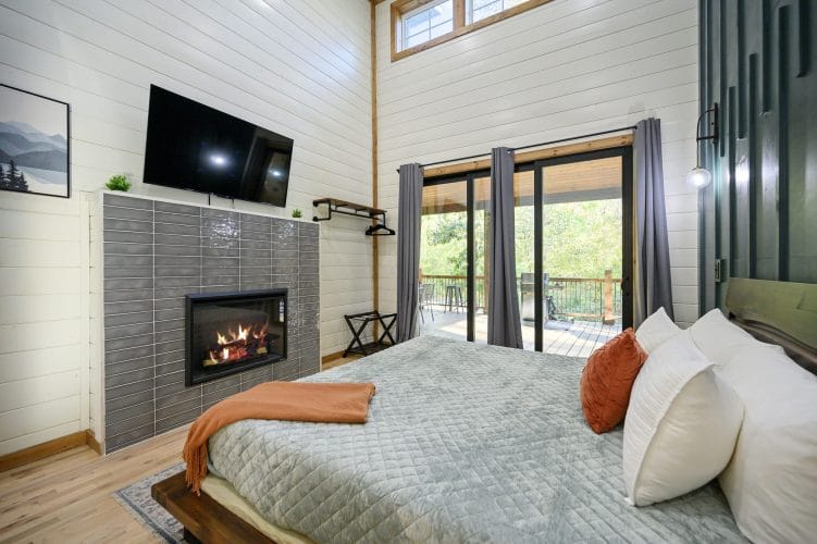 Same bedroom with a gas fireplace in a gray tile wall under the TV, and sliding doors to the deck