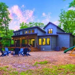 Back of the blue-gray cabin with a deck, fire pit chairs and a playset with a slide on a gravel yard