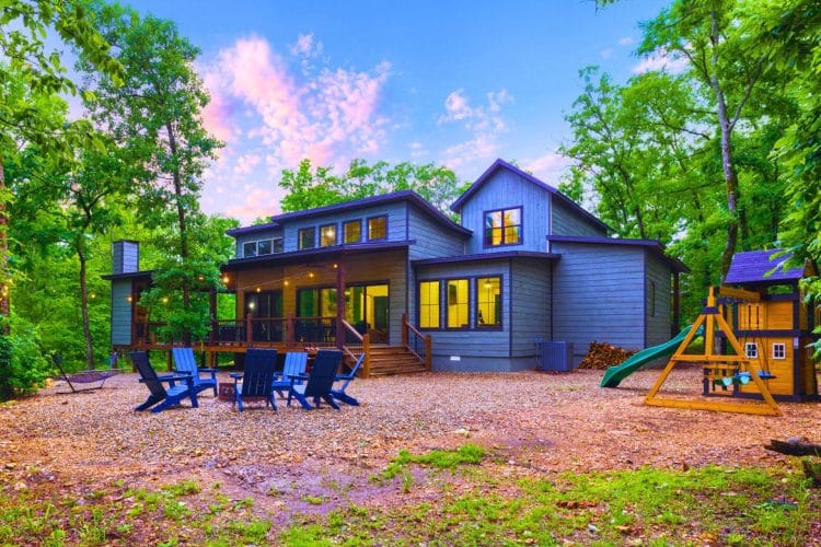 Back of the blue-gray cabin with a deck, fire pit chairs and a playset with a slide on a gravel yard