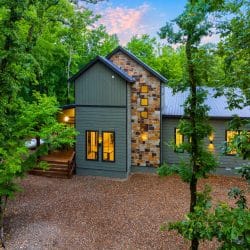 Front of the green-gray cabin with a stone-clad tower and a gravel drive at dusk