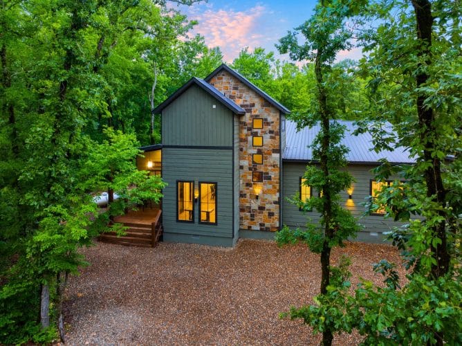 Front of the green-gray cabin with a stone-clad tower and a gravel drive at dusk