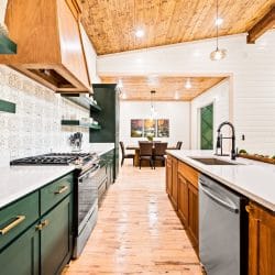 Long kitchen with green cabinets, a wood range hood and patterned tile backsplash