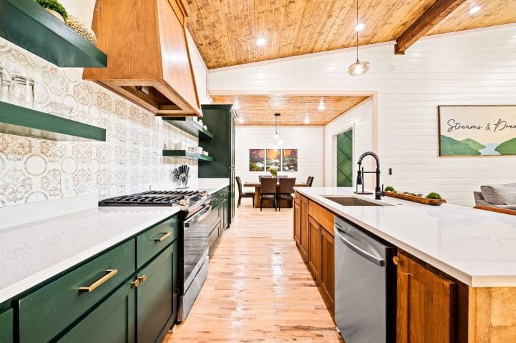 Long kitchen with green cabinets, a wood range hood and patterned tile backsplash