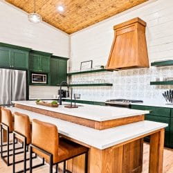 Island with leather stools, green cabinets and a wood range hood