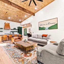 Great room with a gas fireplace, gray sofas and a wood coffee table, the kitchen beyond under a pine ceiling
