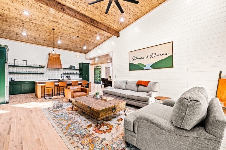 Great room with a gas fireplace, gray sofas and a wood coffee table, the kitchen beyond under a pine ceiling