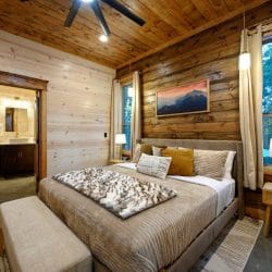 King bed with tan bedding and a bench at the foot, bathroom doorway at left