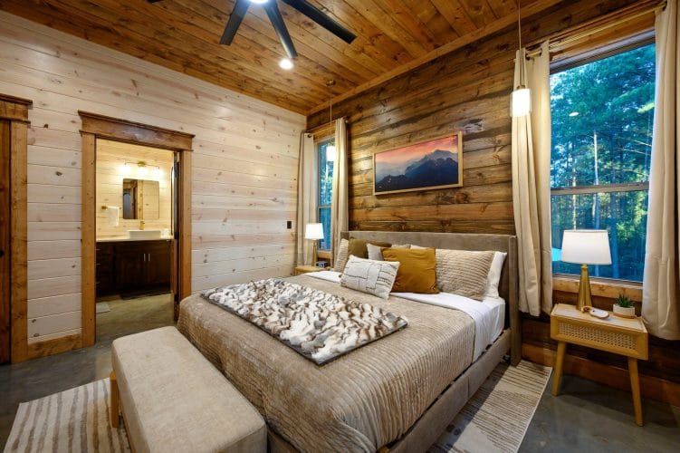 King bed with tan bedding and a bench at the foot, bathroom doorway at left