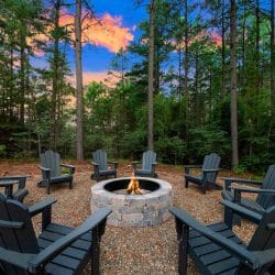 Stone fire pit ringed by dark Adirondack chairs in the pines