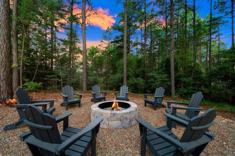 Stone fire pit ringed by dark Adirondack chairs in the pines