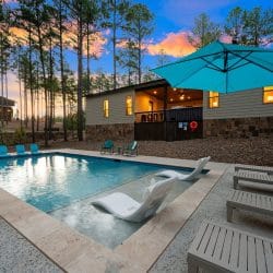 Pool with a teal umbrella and loungers, house with lit porch behind at dusk