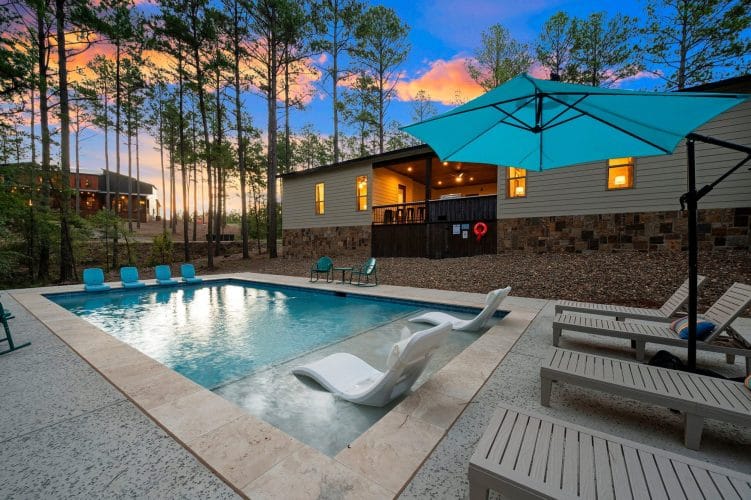 Pool with a teal umbrella and loungers, house with lit porch behind at dusk