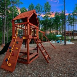 Red wood playset with a climbing wall and slide among the pines, pool behind