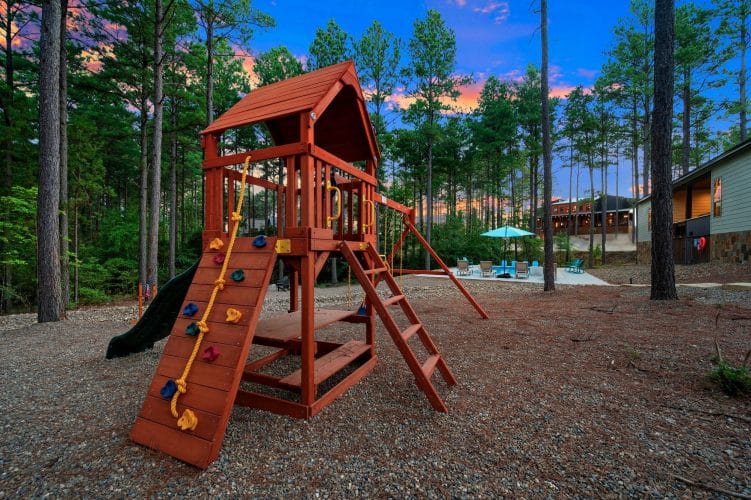Red wood playset with a climbing wall and slide among the pines, pool behind