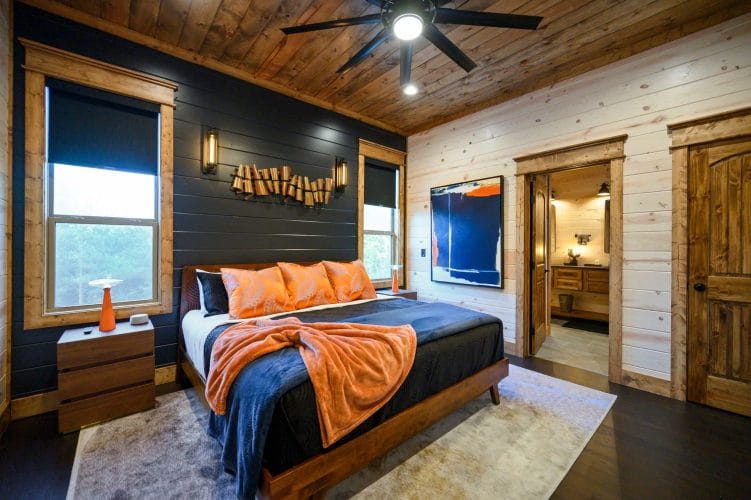 Bed with orange pillows and a blue blanket in front of a dark shiplap wall and a window