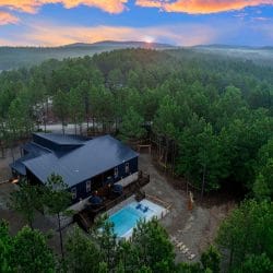 Aerial view of the dark metal roof and cabin edge among tall pines, with a string-lit path in the trees
