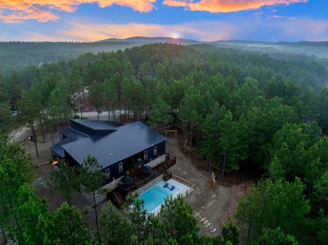 Aerial view of the dark metal roof and cabin edge among tall pines, with a string-lit path in the trees