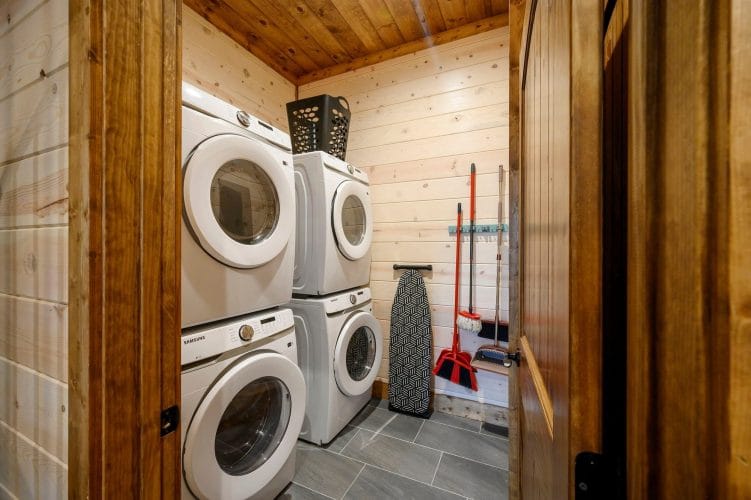 Stacked washer and dryer with an ironing board and mop on the wall
