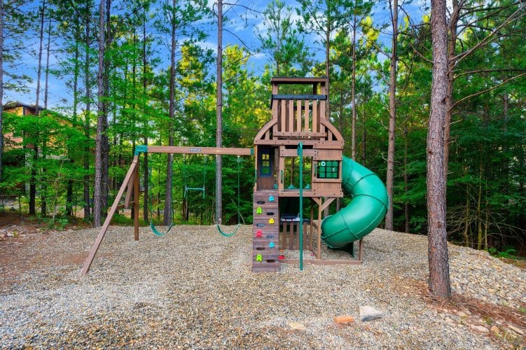 Wooden playset with climbing wall, swings and a green slide in the trees