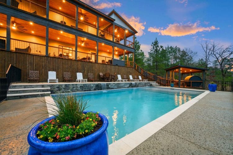 Multi-level lodge with stacked decks lit up at dusk, with the pool in the foreground