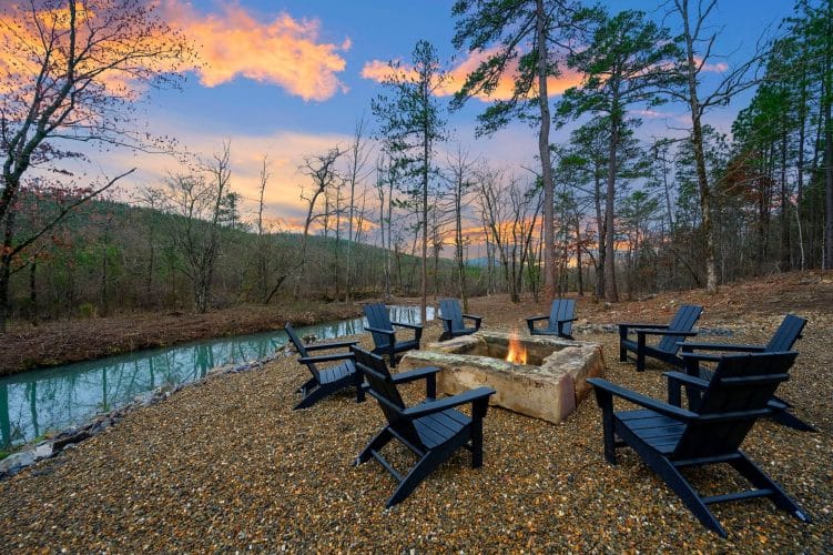 Stone fire pit with black Adirondack chairs on the bank of the creek