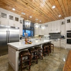 White kitchen island with five leather stools, stainless refrigerator and pendant lights