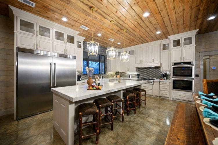 White kitchen island with five leather stools, stainless refrigerator and pendant lights