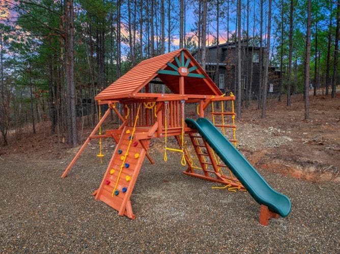 Wooden playset with a green slide and climbing wall in the pines