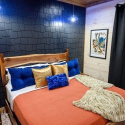 King bed with coral cover and blue and gold pillows against a navy shingle wall
