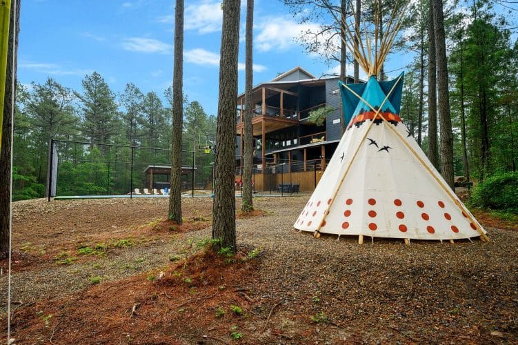 Teepee in the pines with the house behind it