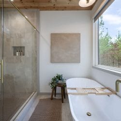 Freestanding soaking tub next to a glass shower, with a window onto the pines