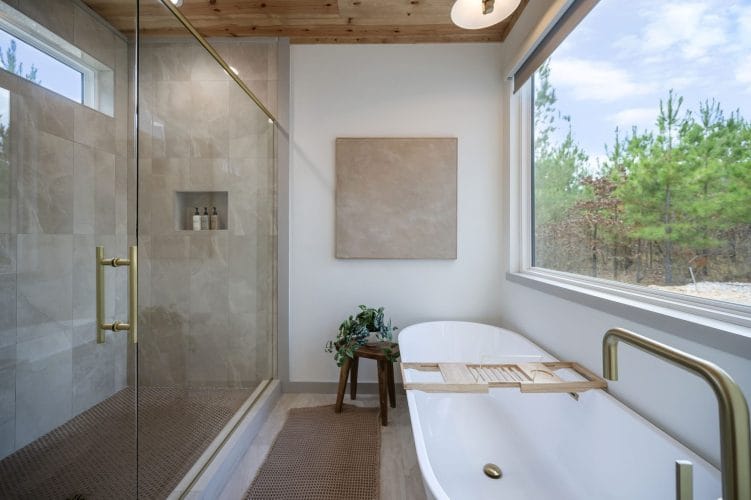 Freestanding soaking tub next to a glass shower, with a window onto the pines