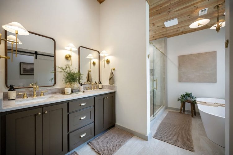 Double vanity with dark cabinets, brass fixtures, glass shower and tub in the background