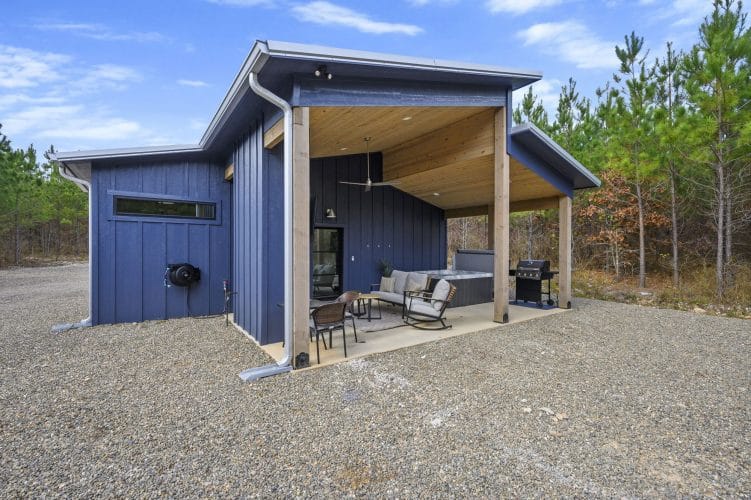 Side of the cabin with a covered patio, sofa, hot tub and grill on a gravel lot