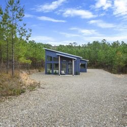 Cabin seen from far down the gravel lot, surrounded by pine woods