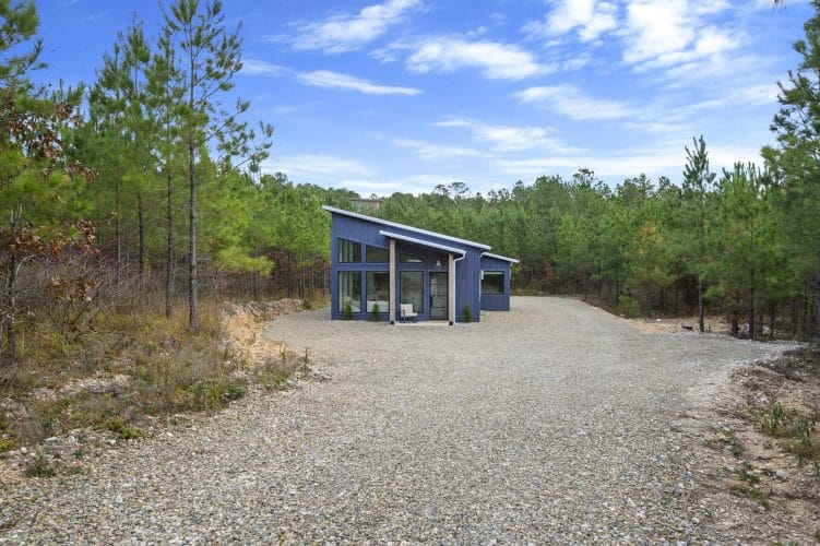 Cabin seen from far down the gravel lot, surrounded by pine woods