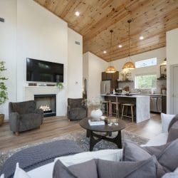 Open living room with a cream sofa, two gray armchairs and a fireplace under a TV, wood plank vaulted ceiling and the kitchen beyond
