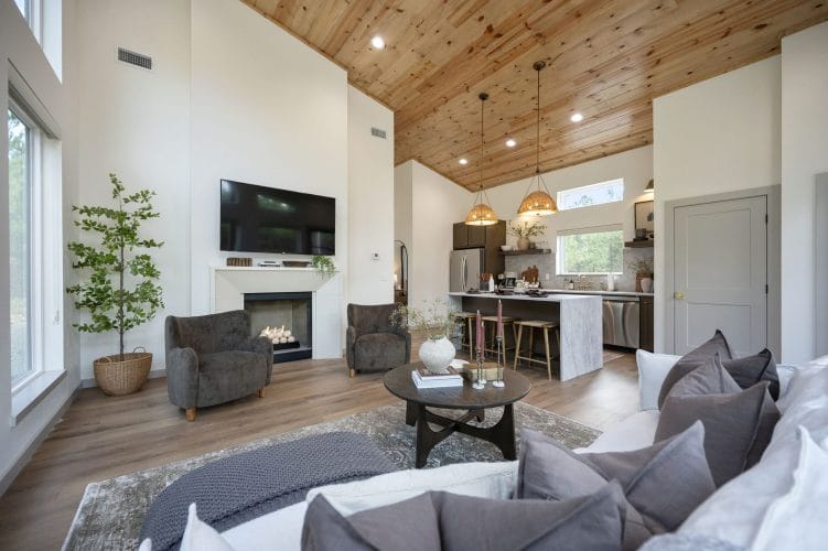 Open living room with a cream sofa, two gray armchairs and a fireplace under a TV, wood plank vaulted ceiling and the kitchen beyond