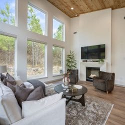 Living room windows on two walls looking onto pines, sofa and fireplace at the far end